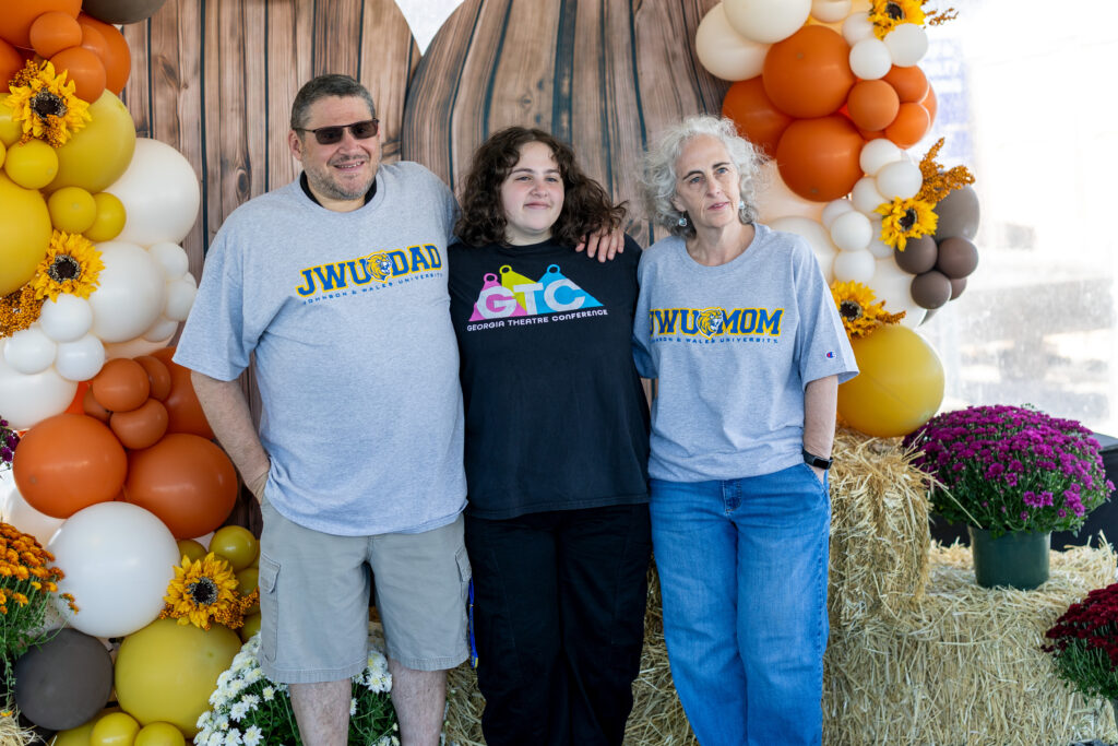 Three people pose together in front of a festive backdrop of balloons and hay bales, wearing themed shirts for Johnson 