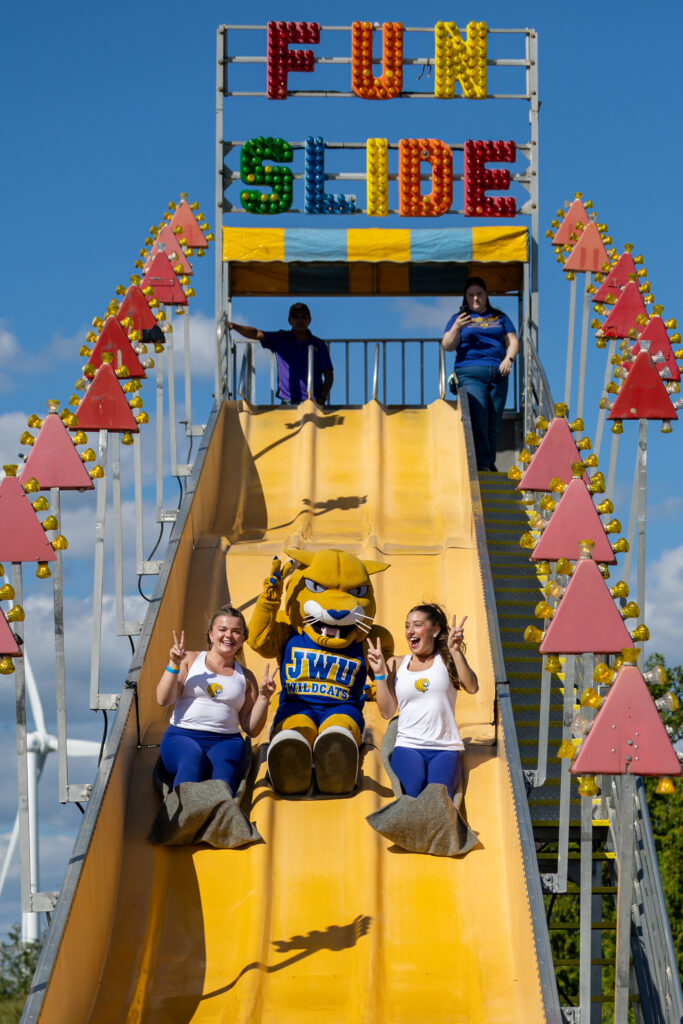 A mascot and two people slide down a bright yellow slide at a fair, with a large "FUN SLIDE" sign overhead.