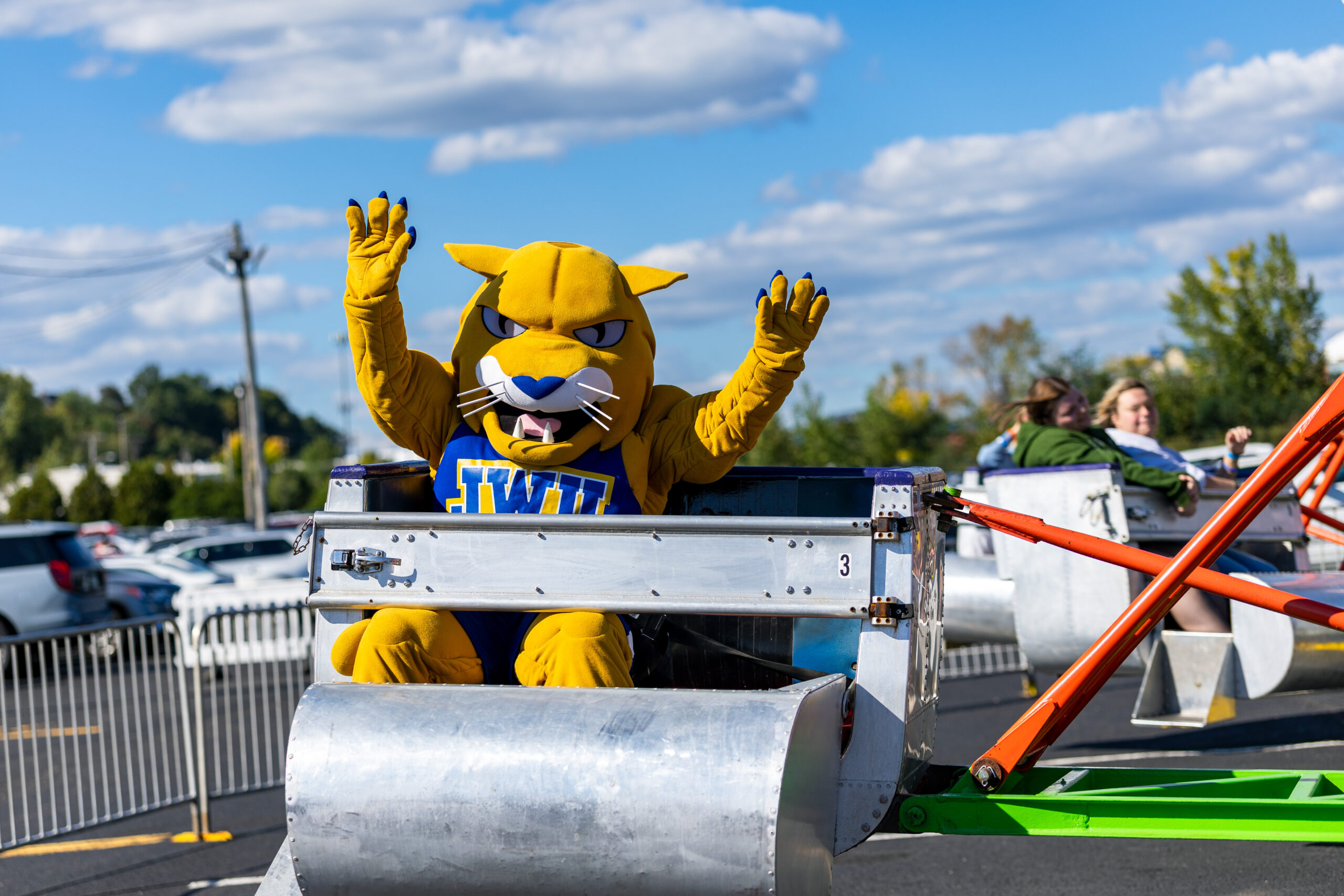 A yellow mascot with blue claws and a blue jersey with "JWU" on it waves enthusiastically from a metal ride vehicle under a bright blue sky.