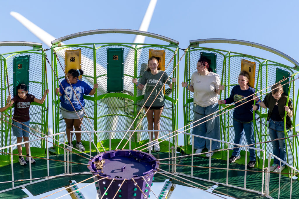 Six individuals stand on a green, circular platform with numbered compartments, holding onto ropes as a large wind turbine spins in the background under a clear blue sky.