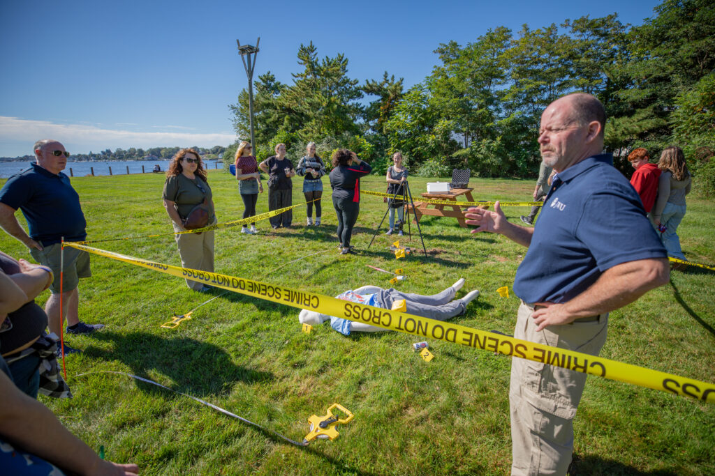 A group gathers outdoors behind crime scene tape as an instructor demonstrates a forensic investigation using a mannequin and evidence markers.