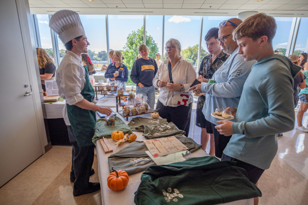 A chef in a tall hat and apron serves food and merchandise to a group of people at an indoor event with large windows.