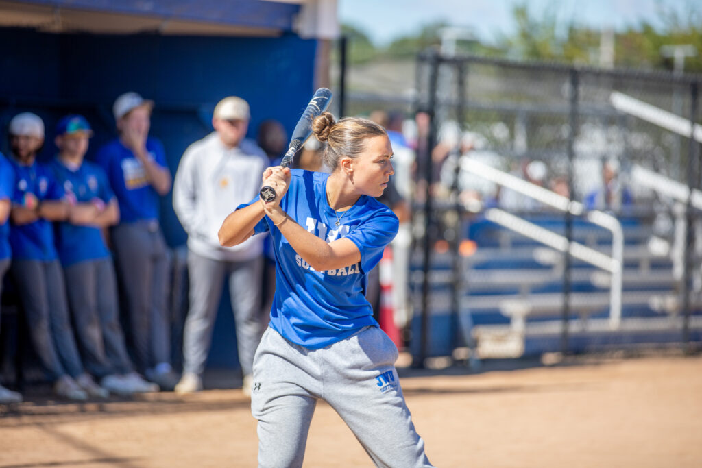 A softball player in a blue jersey and gray pants stands at bat, gripping a bat and preparing to swing, with teammates and spectators blurred in the background.