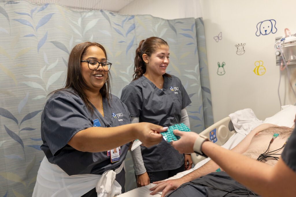 Medical staff in scrubs assist a patient in a hospital room, with one person handing something to another while others observe.