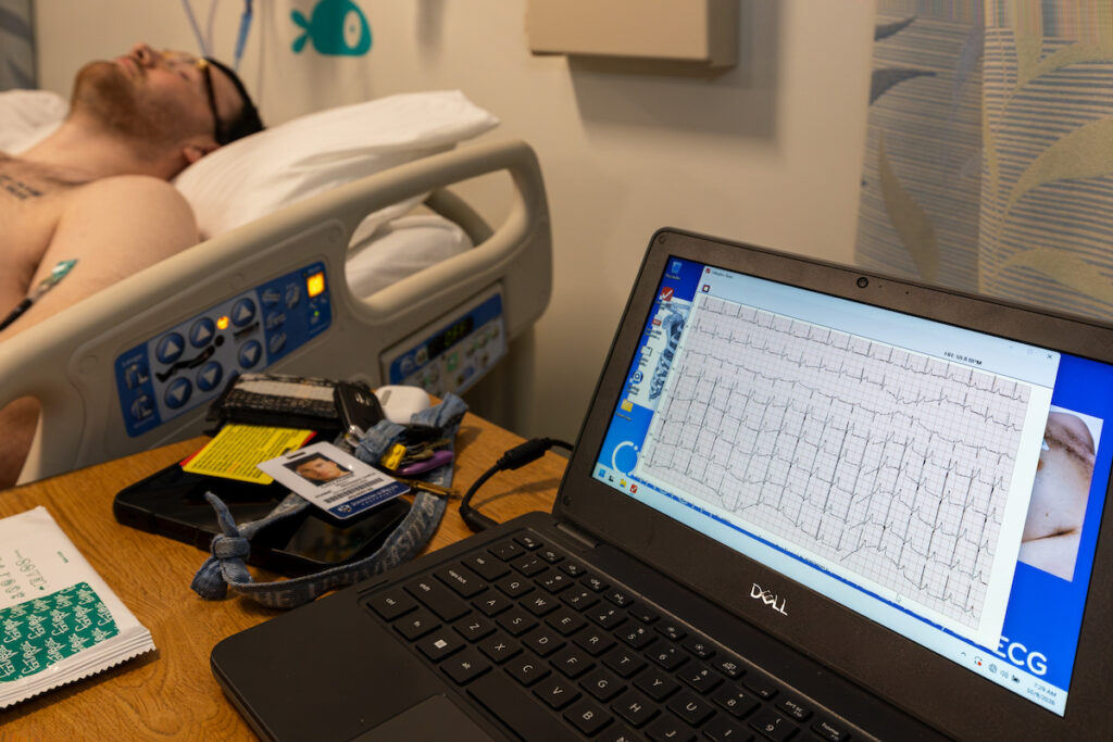 A person rests in a hospital bed while a laptop displays an ECG reading on a wooden table beside them.