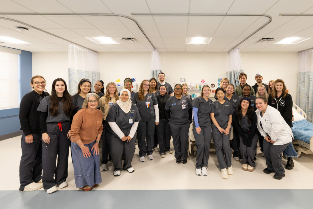 A diverse group of healthcare professionals poses together in a hospital room, wearing scrubs and medical attire.
