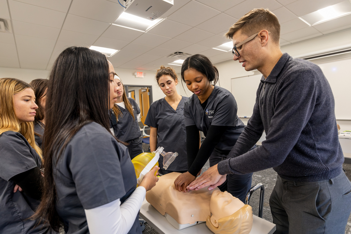 A group of students in scrubs gathers around a medical training mannequin as one student demonstrates CPR techniques while others observe.