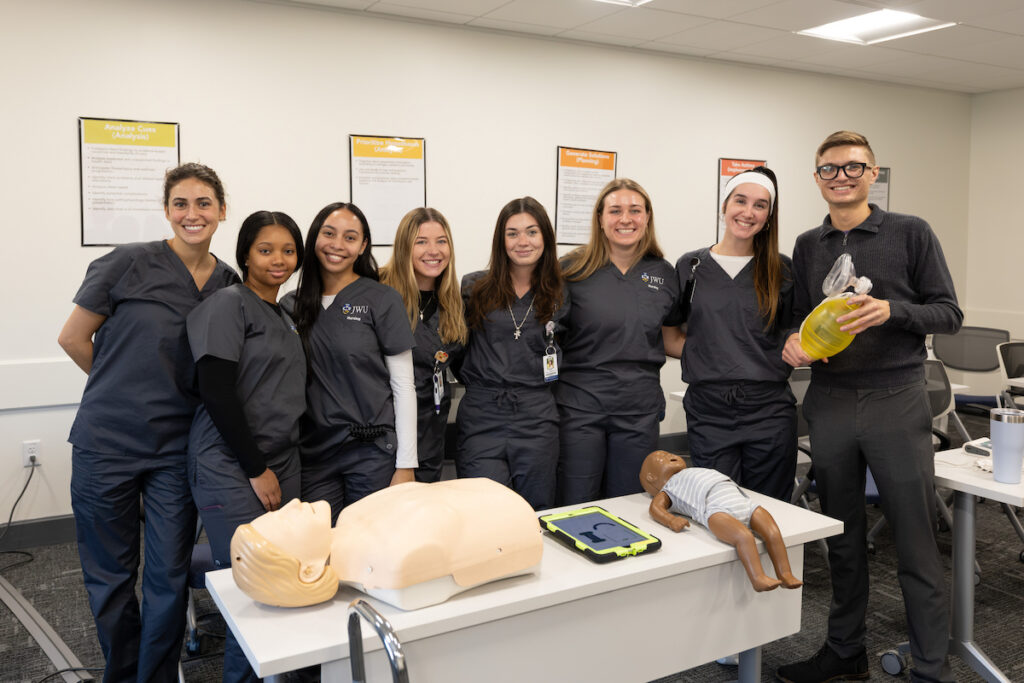 A group of medical students in scrubs pose with training mannequins and equipment in a classroom setting.
