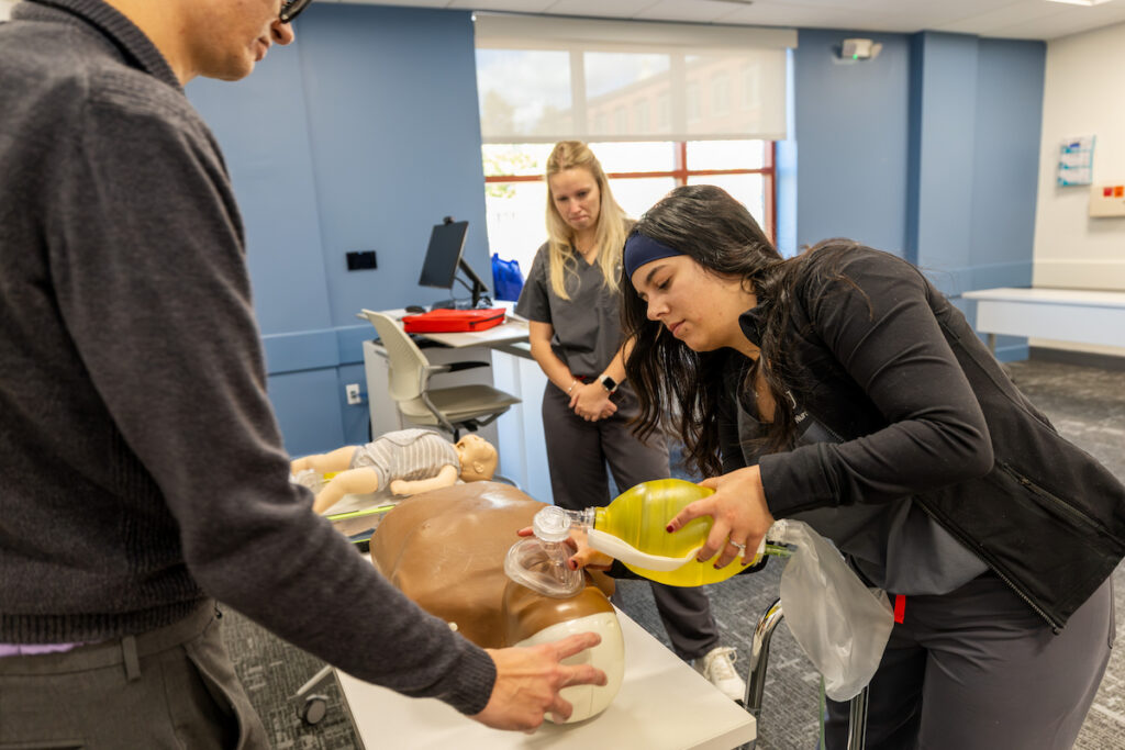 A person performs CPR on a mannequin while another observes, in a clinical training setting.