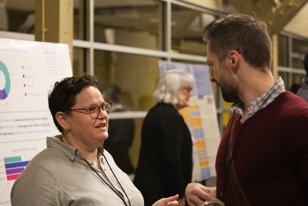 Two individuals engage in conversation at what appears to be an academic or professional conference, with presentation boards visible in the background.