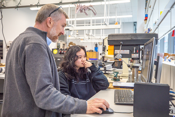 Two individuals collaborate in a workshop, focused on a computer screen with machinery visible in the background.