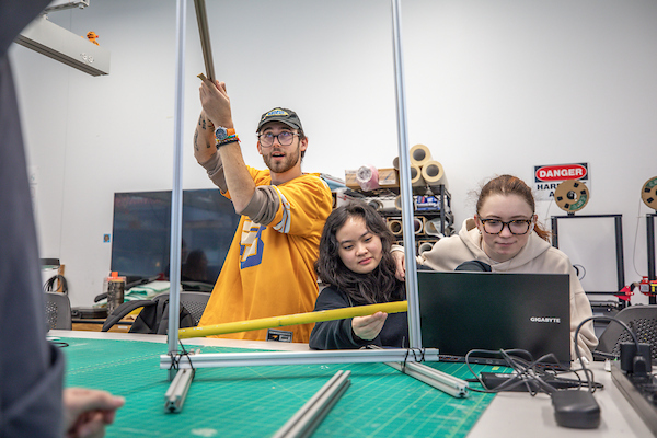 Three individuals collaborate in a workshop setting, with one holding a long metal rod while another works on a laptop and the third observes closely.