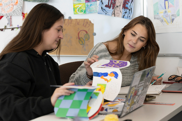 Two individuals collaborate on a creative project in a classroom setting, surrounded by colorful artwork and school supplies.