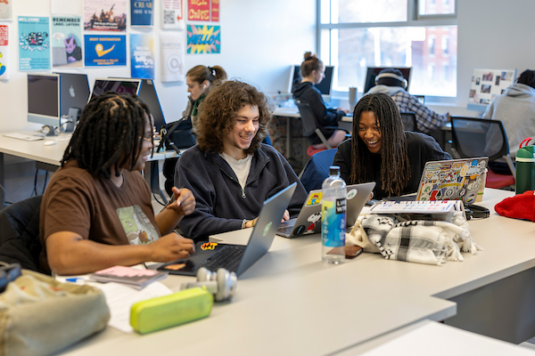 Three individuals laugh together while working on laptops at a shared desk in a bright, busy office space.