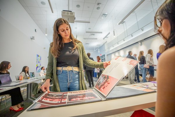 A person examines promotional materials at an event in a modern, well-lit indoor space with other attendees and tables in the background.