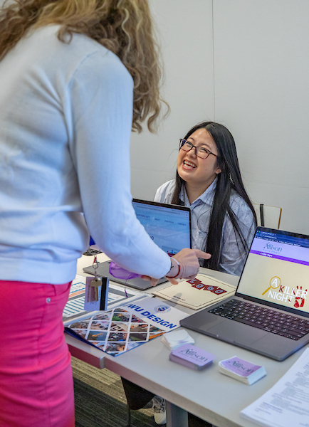 A person in a light blue top and pink pants gestures toward a smiling individual seated at a desk with laptops and design materials.