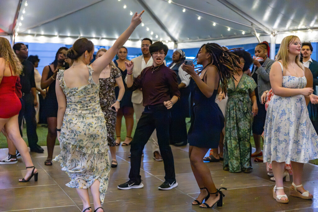 A group of people dance energetically under a tent adorned with string lights.