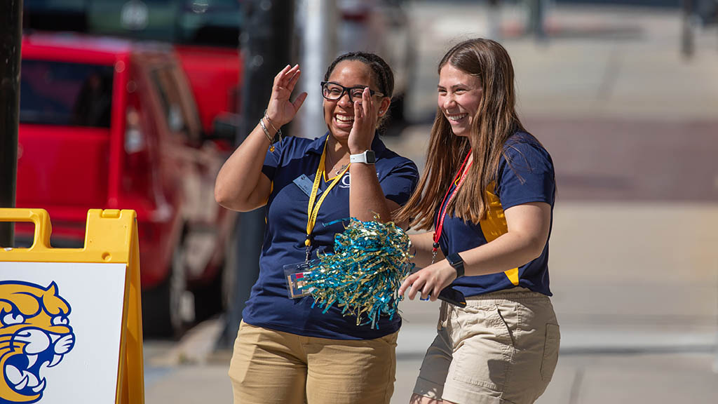 two young women wearing JWU CAT shirts excitedly greet drivers on the side of the road
