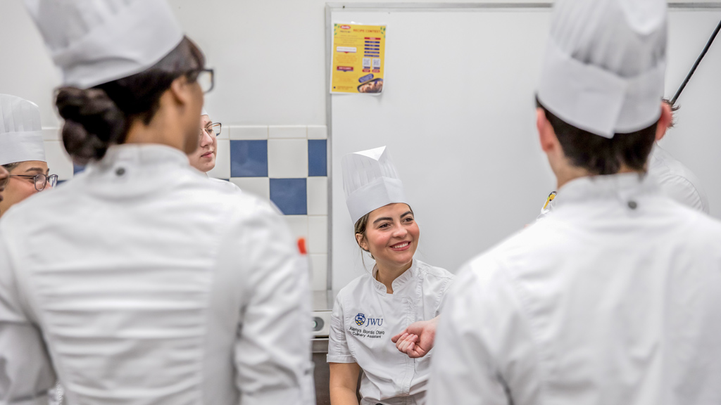 a photo showing one student's face in focus and the backs of other students gathered in chef's whites in a JWU kitchen