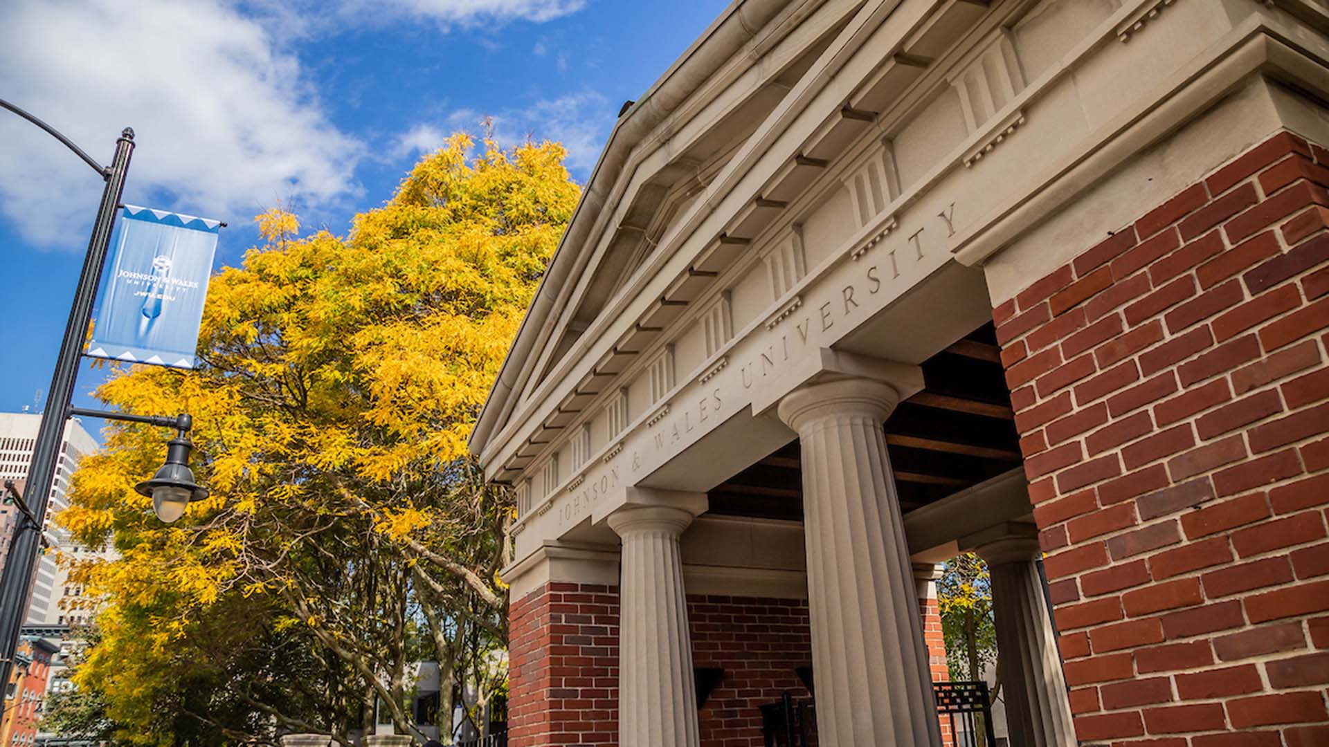 Entrance of Johnson u0026 Wales University with a brick structure featuring classical columns, under a clear sky with a vibrant yellow tree nearby.