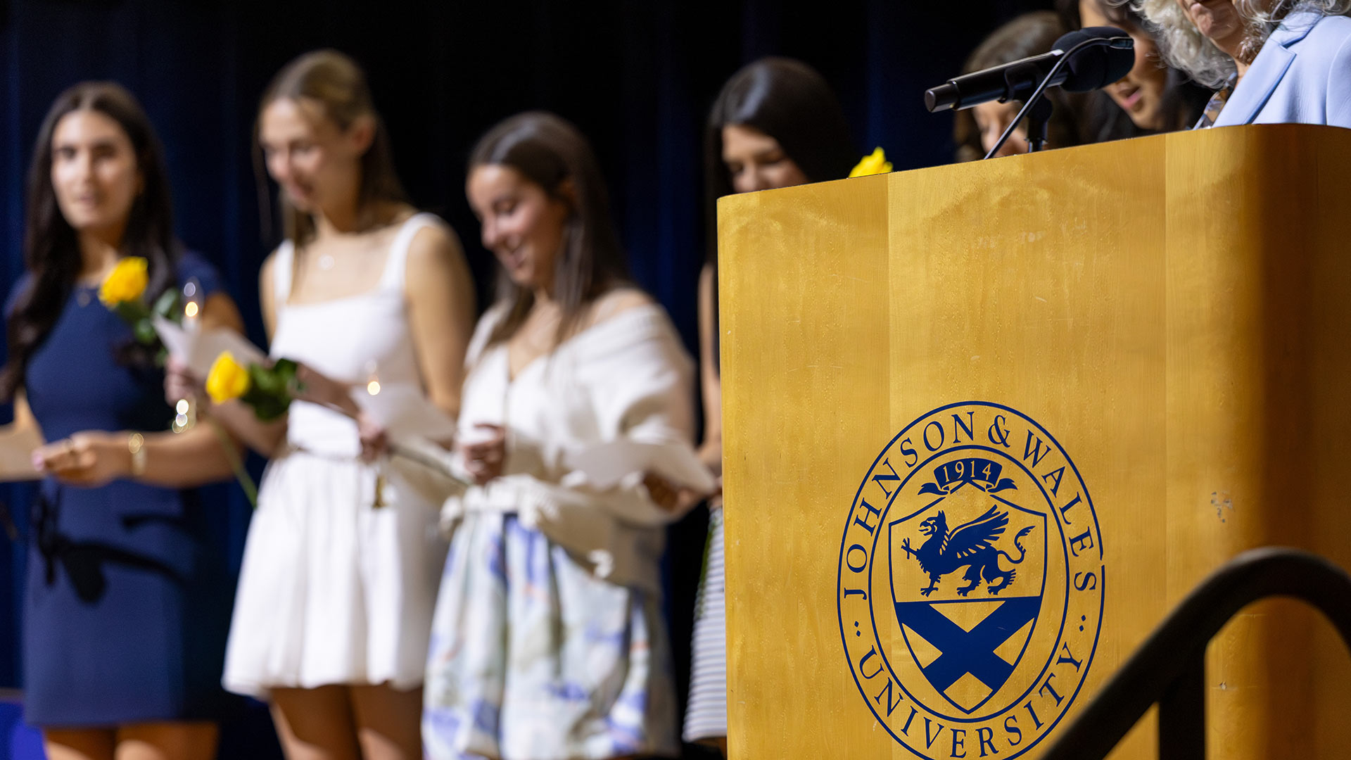 a JWU logo on a podium is in focus while OTD students take an oath in the background